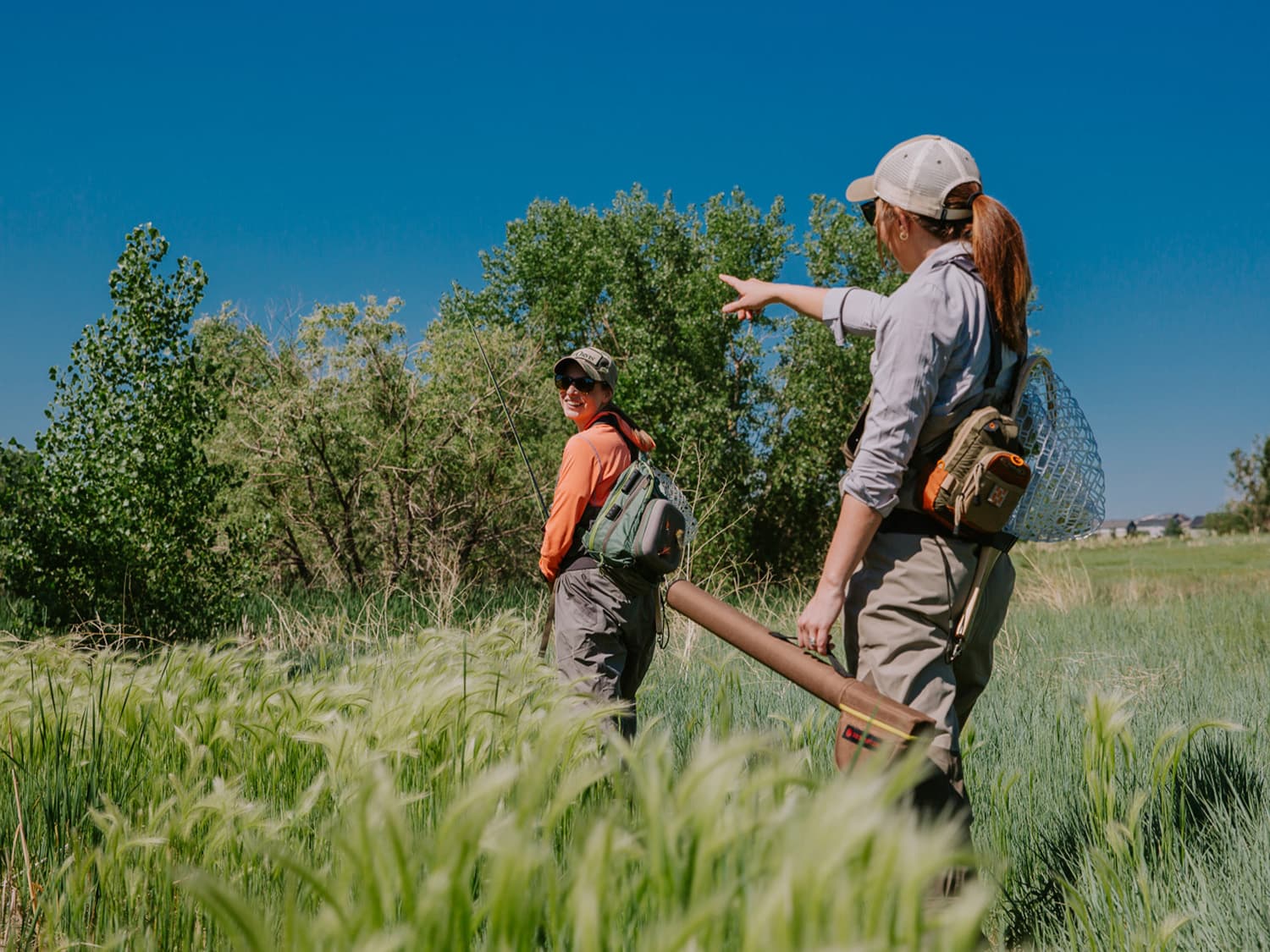 Two people hike through knee-high grass carrying fly-fishing gear.
