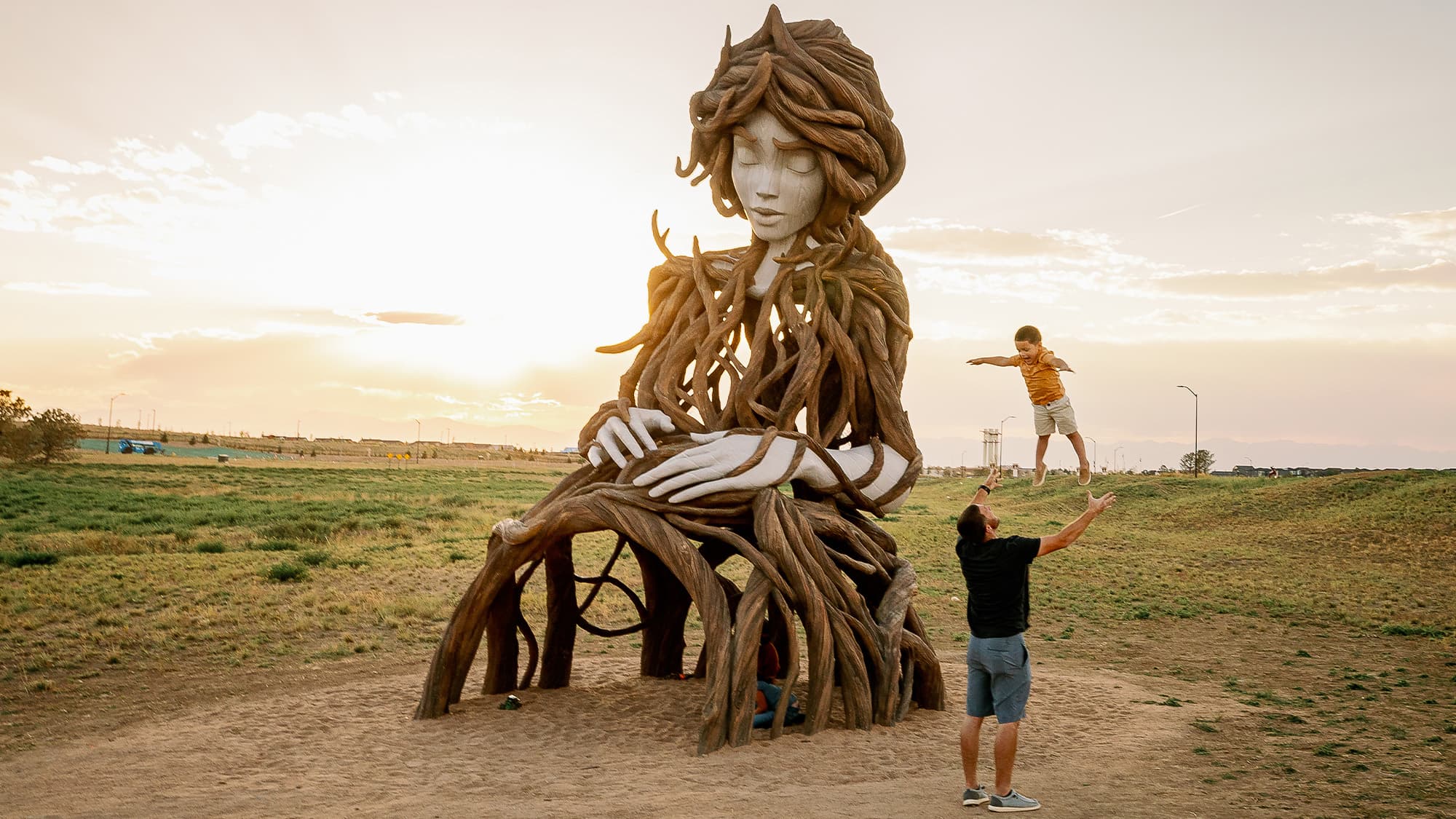 A person throws a child up in the air and catches them in a lush park next to Umi, a sculpture that blends a woman with a tree