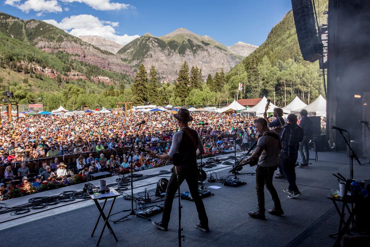 A band plays on stage and looks out to a crowd and mountain scene