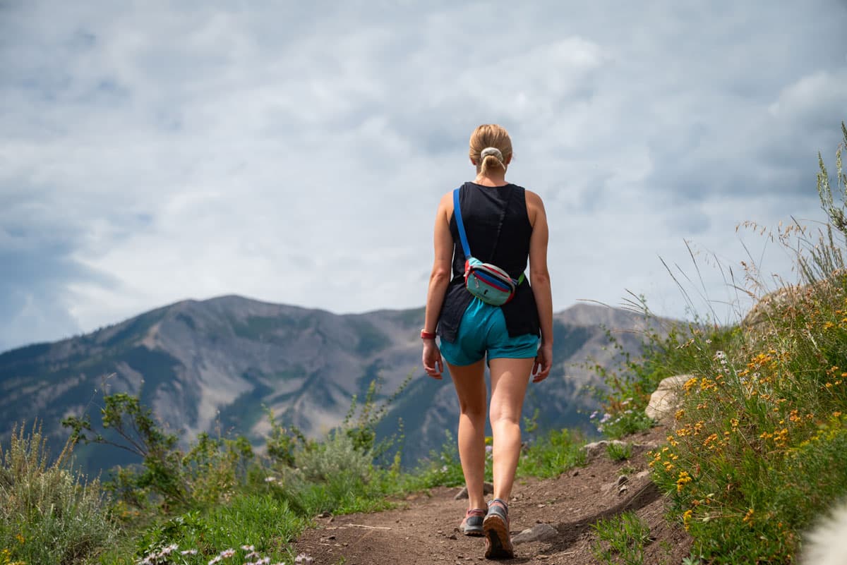 Woman hiking a trail past wildflowers with mountains in the background.
