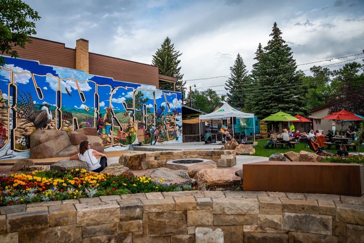 People sitting at tables listening to live music under colorful tents at a park event.