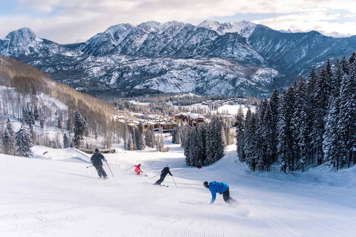 Skiers and snowboarders carving fresh powder downhill with sweeping mountain views in the background.