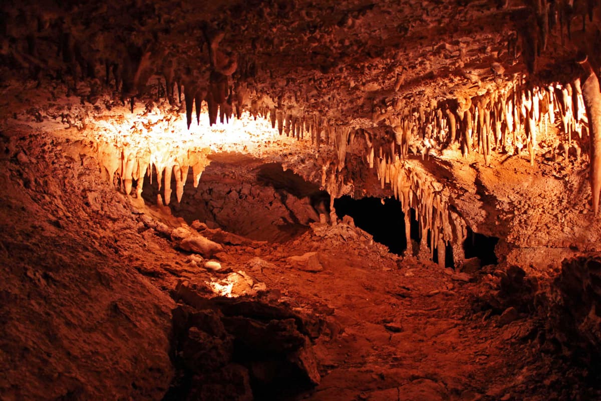 Lights shine on the jagged, toothy stalactites growing from the roof of a Colorado Springs' cave. The cave looks deep and the walls have burnt orange-red color.