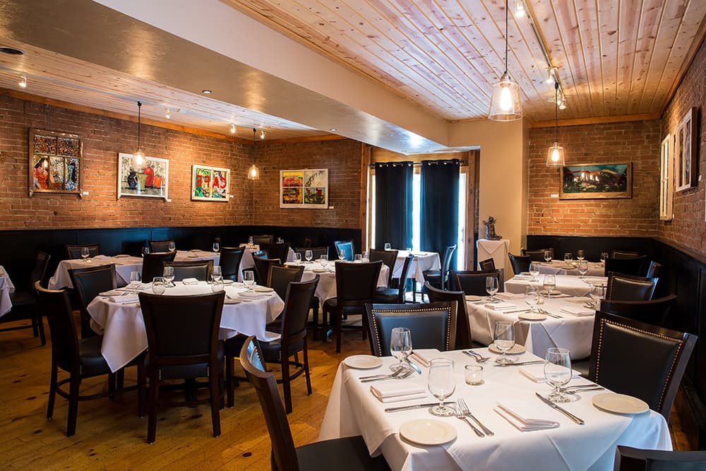 The empty dining room waits for diners. Several white-tableclothed tables are set with wine glasses, white plate and silverewear. Brown leather chairs sit empty. A series of paintings hang on the brick walls.