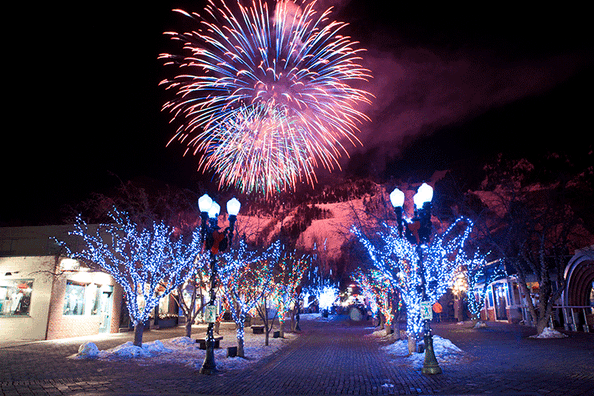 Fireworks explode in a night sky above trees lit with white lights