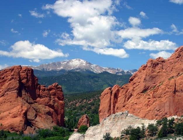 Two red-rock formations sit on the sides of the image with a window to the snow-capped Pikes Peak with green rolling hills below it and a blue sky with white clouds.
