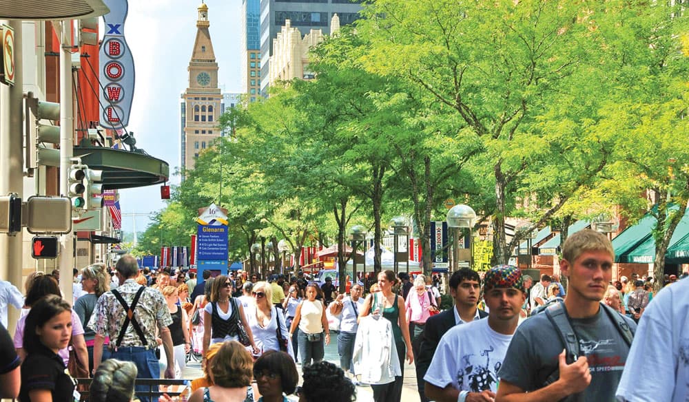People walk down the street of Denver's 16th Street Mall on a sunny day with a bright green tree in the background