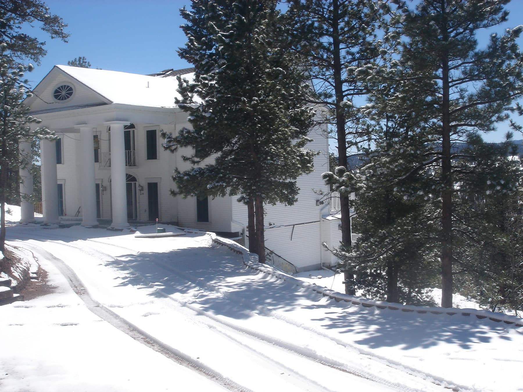 A white building with four thick, roman columns stands among towering pines in Woodland Park, Colorado. Everything is covered in a soft blanket of snow.