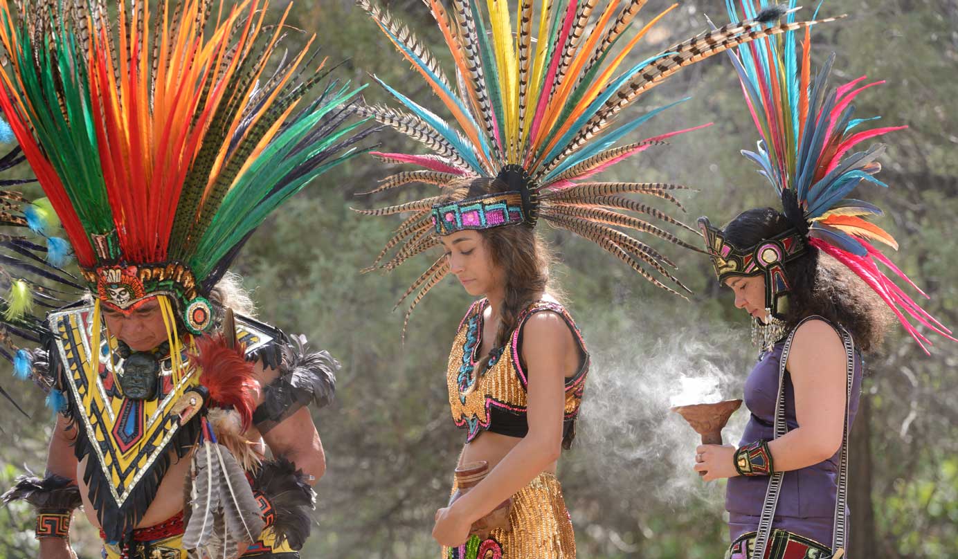 Three Native American people wearing brightly colored headresses made of brilliant feathers and beautiful beadwork walk with their heads bowed