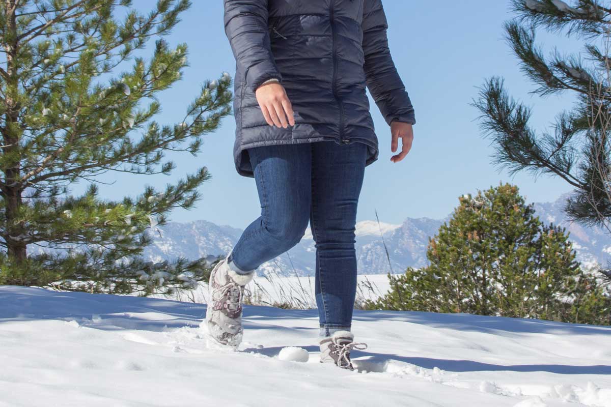 A person walks through ankle-deep snow wearing off-white winter shoes. Small shrubs and the Colorado Rocky Mountain are in the background.