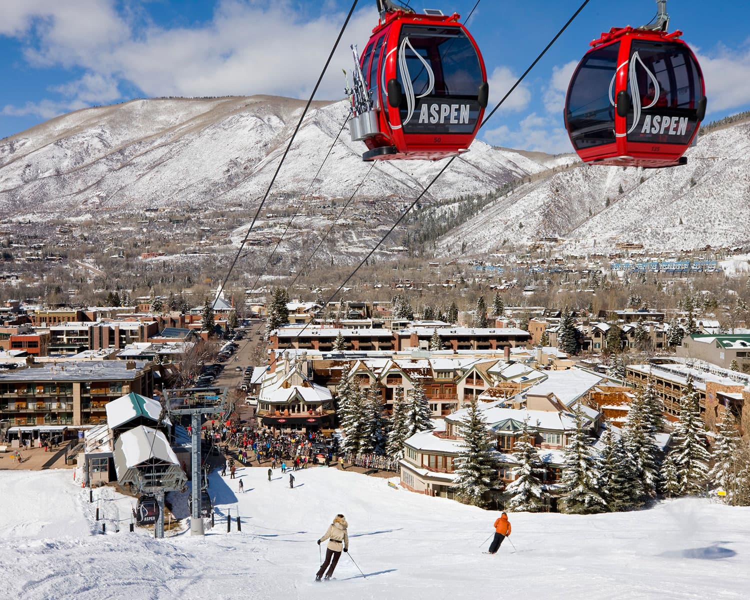Two red and black Aspen Snowmass gondolas dangle over the ski area with hotels, mountains and a bright-blue sky in the background. Everything is covered in a layer of snow.