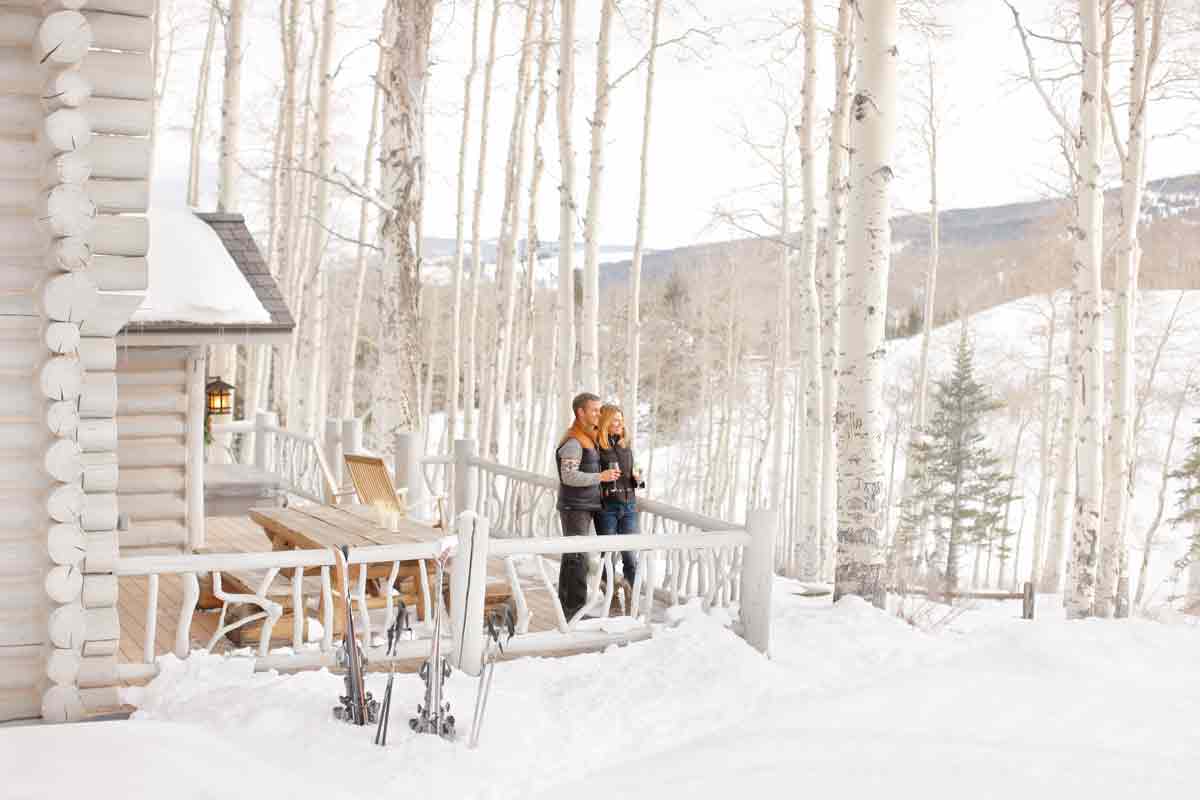 A couple stands close together on an outdoor deck of a cabin surrounded by bare aspens and a thick blanket of snow in Beaver Creek, Colorado.
