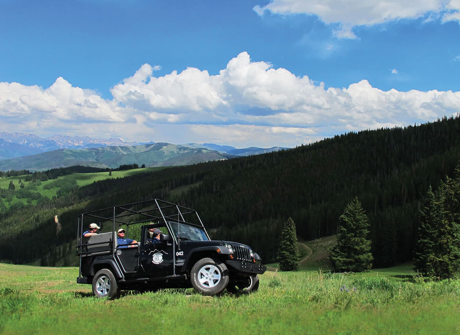 A black, topless Jeep carries passengers and goes off-roading through a grassy meadow surrounded by forested mountainsides near Beaver Creek Resort in Colorado.