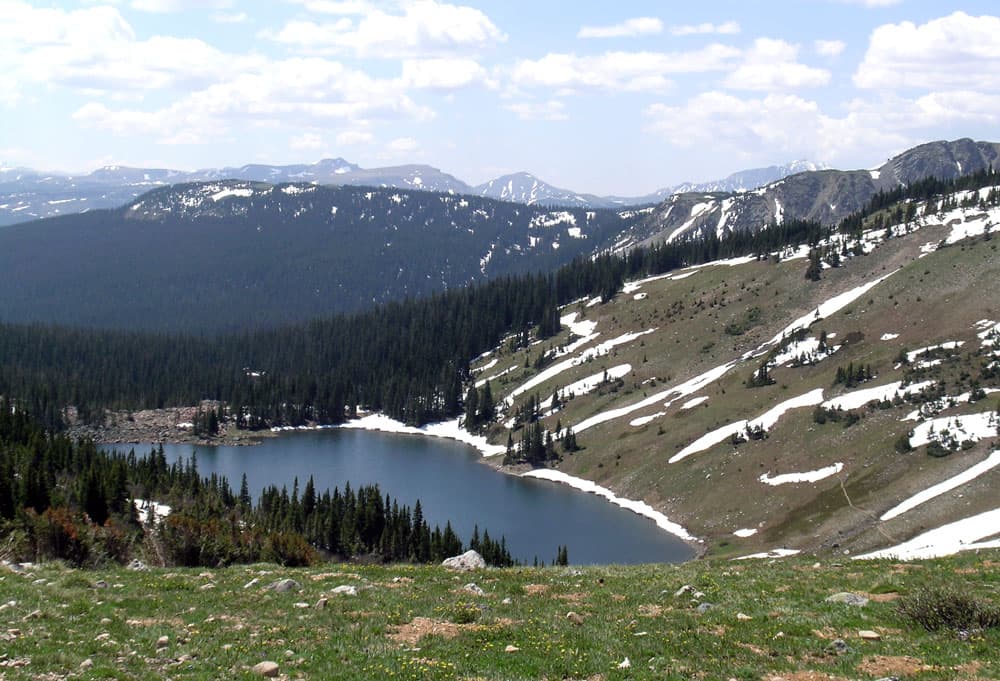 A navy-blue lake near Fort Collings sits calm and placid surrounded on three sides tall evergreen. On the last side is a steep, almost barren mountainside with streaks of spring snow.