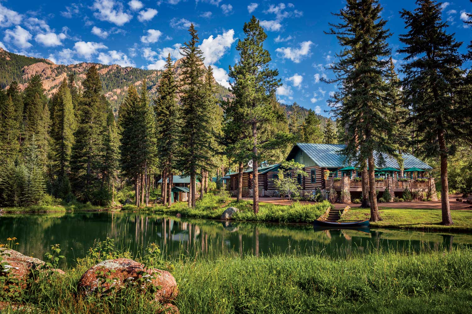 A cozy wood cabin with a faded-blue roof is set next to a pine tree-lined pond in Colorado.  A rugged mountain face watches over the area in the background.