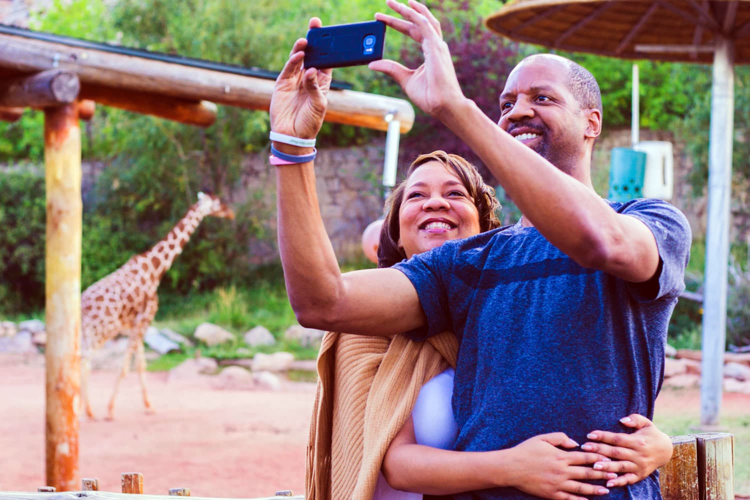 A couple embraces and smiles as they pose for a selfie at the giraffe exhibit during a VIP tour of Cheyenne Mountain Zoo in Colorado Springs.