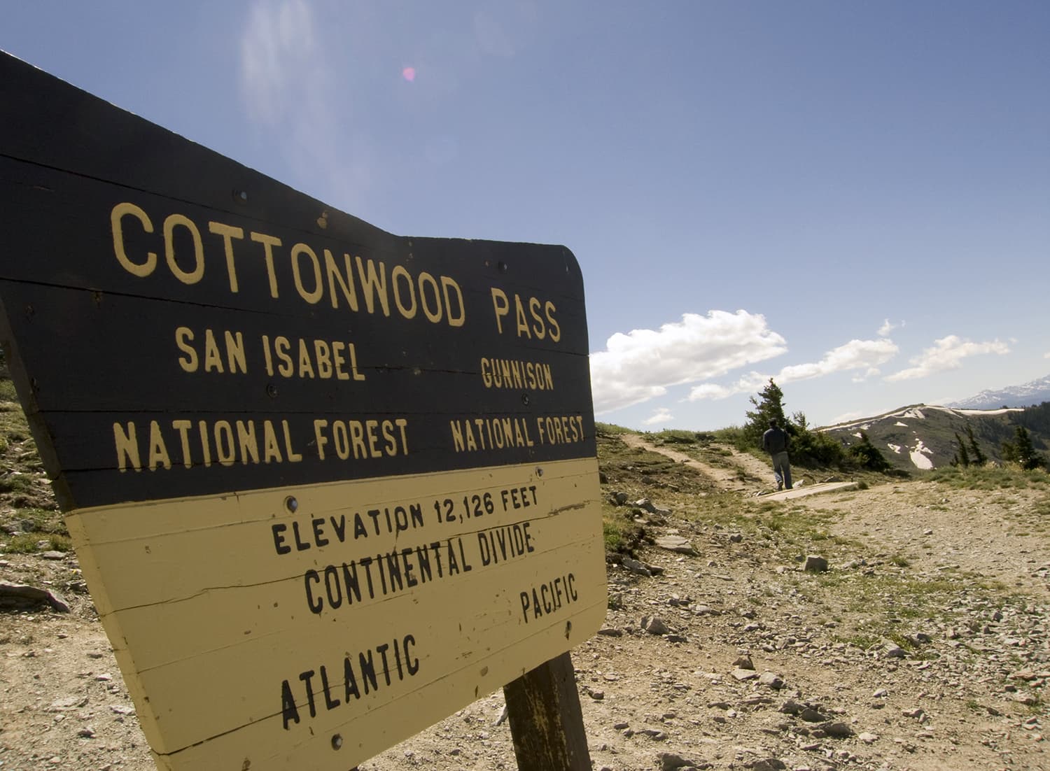 A painted wood sign at Cottonwood Pass in Colorado gives details about the Continental Divide. To the side a hiker stands on the trail and admire the mountains in the distance.