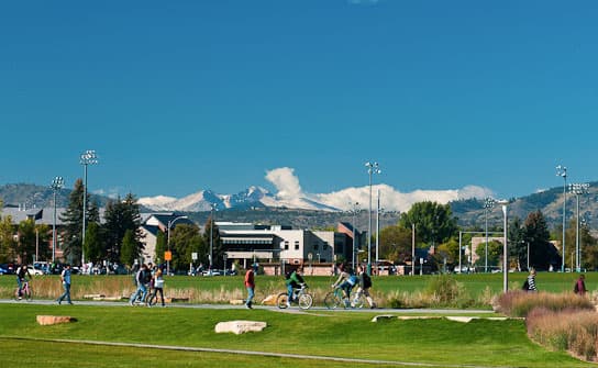 Students bike and walk on a Colorado State University campus path. In the background are university buildings and snow-covered peaks.