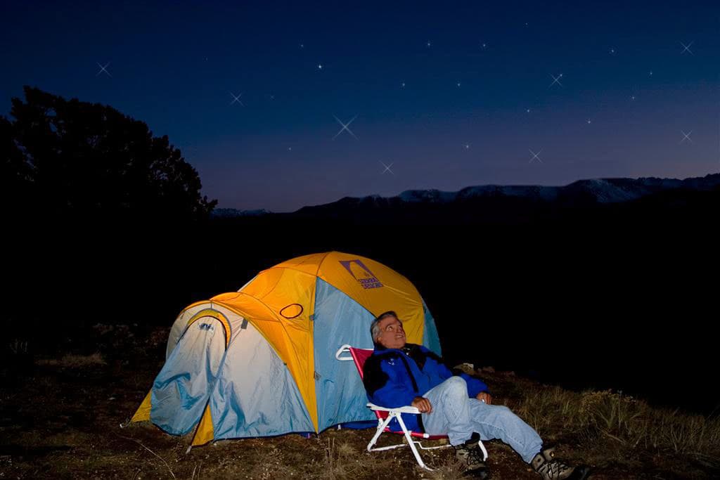 A man in a camp chair gazes up at the stars in front of his orange and blue tent, which glows from an lantern inside