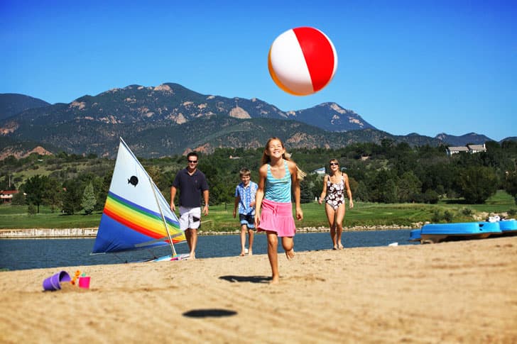 With a lake and peaks in the backround a family chases after a beach ball on the shore