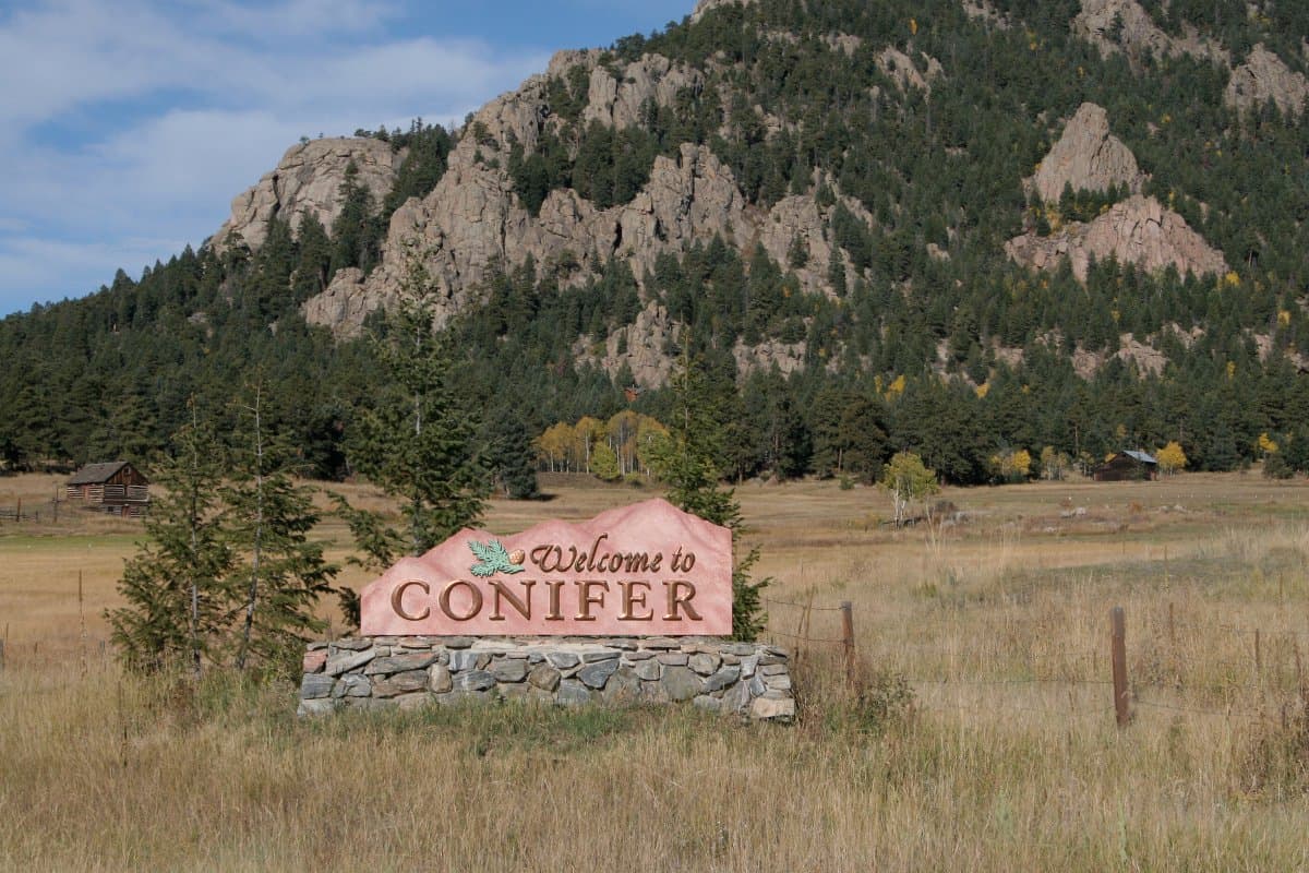 A sign sits in a field of brown grasses with a mountain that's dotted with evergreen trees in the background. The sign, which is made out of a red stone sits atop a grey-stone base and reads "Welcome to Conifer" with a leaf next to the "Welcome."