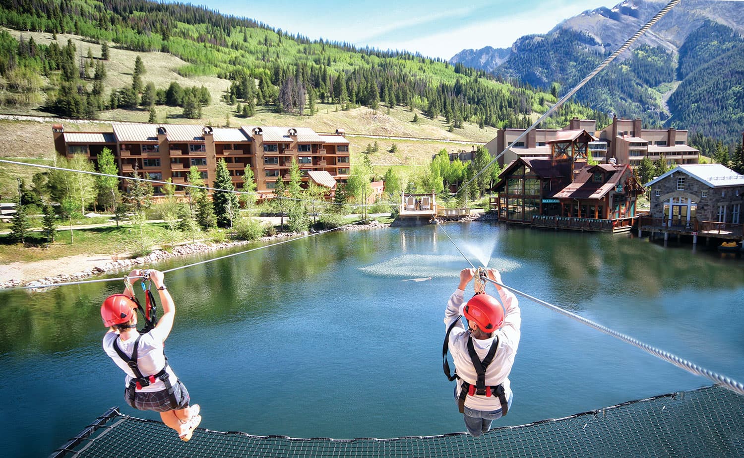 Two adventurers wearing red helmets and strapped into harnesses whiz down a zipline over a lake at Copper Mountain Resort in Colorado.