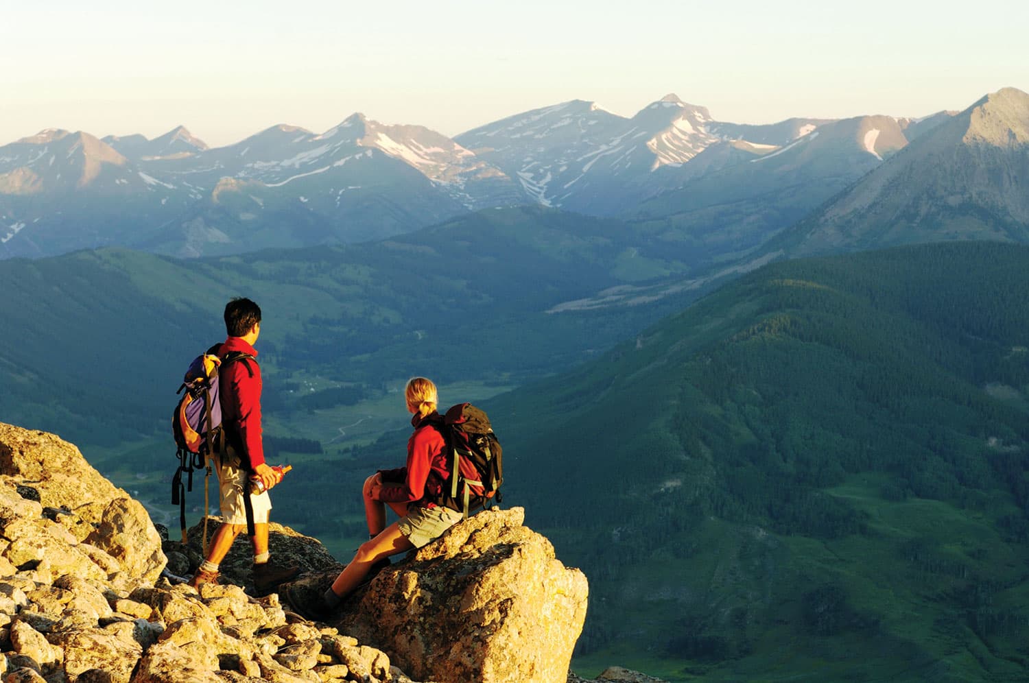 Two hikers in matching red jackets take a break at a rock outcrop near Crested Butte Mountain Resort in Colorado.  They admire the view of the verdant valley and majestic peaks.