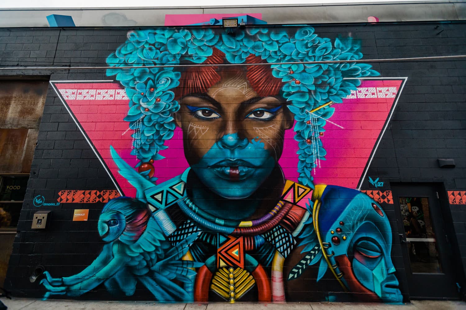 A mural on the side of a black-painted brick building in Denver, Colorado, features an person with vibrant turquoise flowers dripping down from their head.
