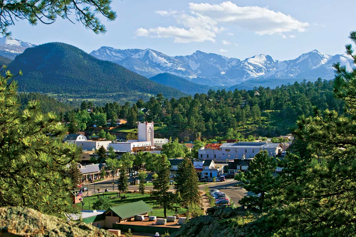A cozy downtown area sits peacefully in the Colorado sun and is surrounded by a thick, lush forest. In the distance are one tree-covered peak and a line of snow-blanketed peaks.