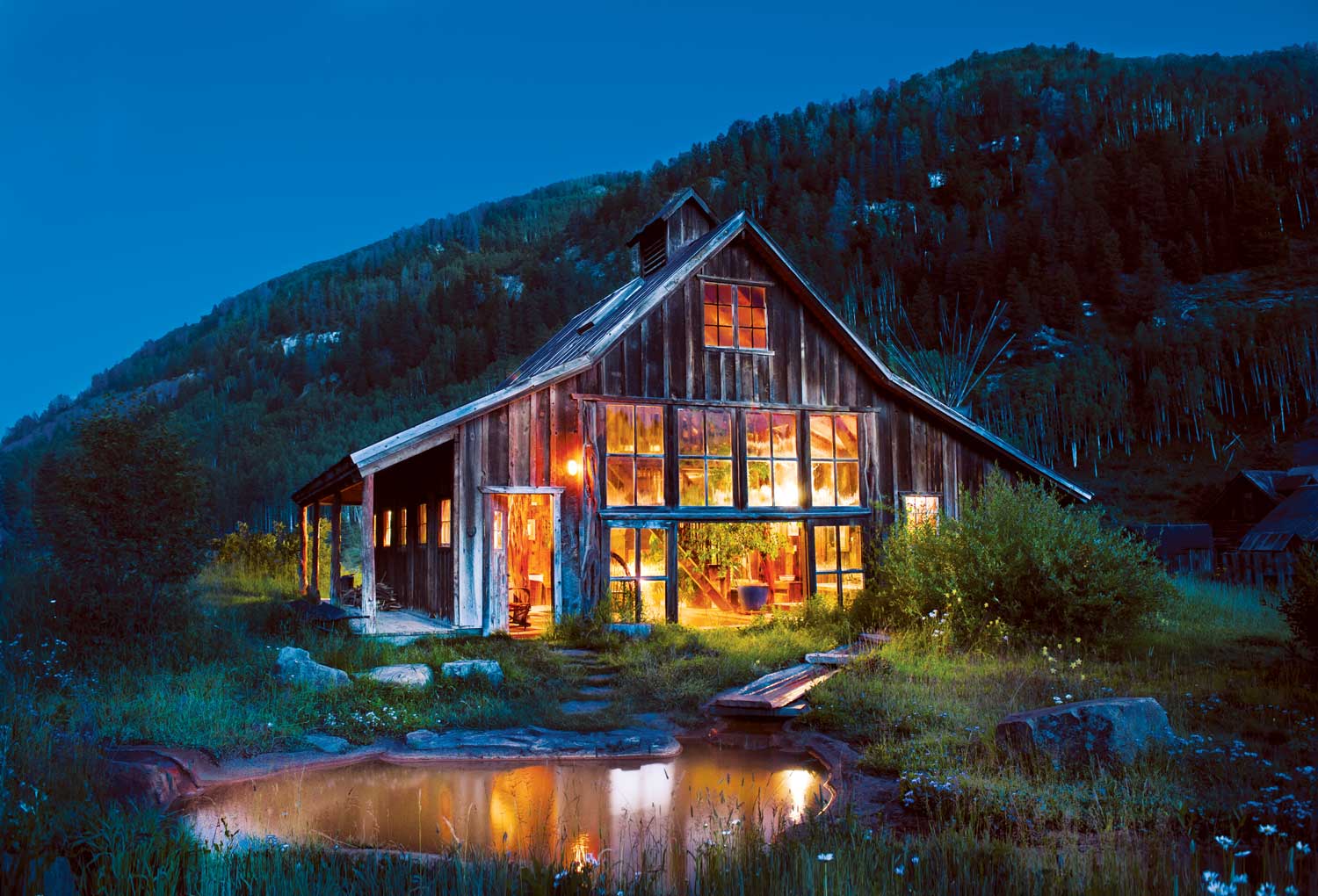 A worn farm building in Colorado glows with golden light against the dark of the evening.  A few stone steps away is a small pool of hot water.