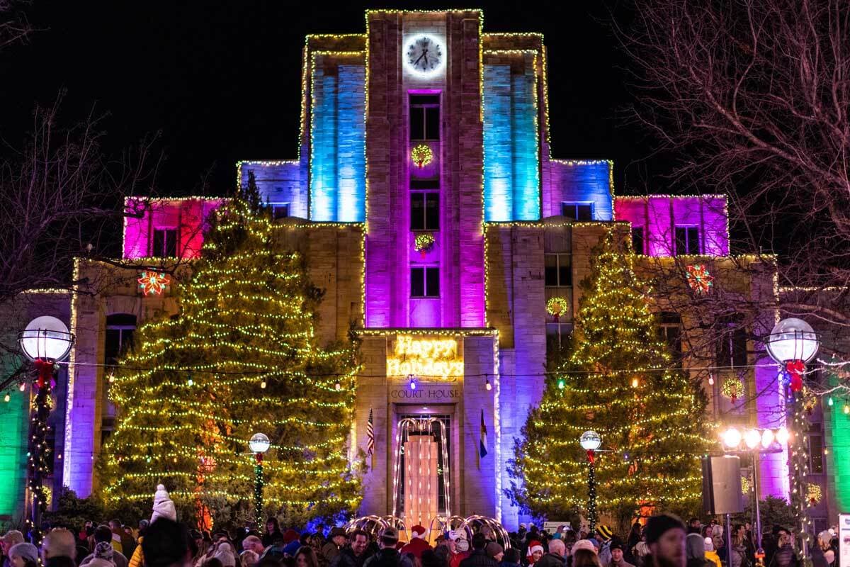 In downtown Boulder, Colorado, two evergreen trees are strung with golden lights. The trees frame the entrance of a building lit with blue, pink, red and purple flood lights.