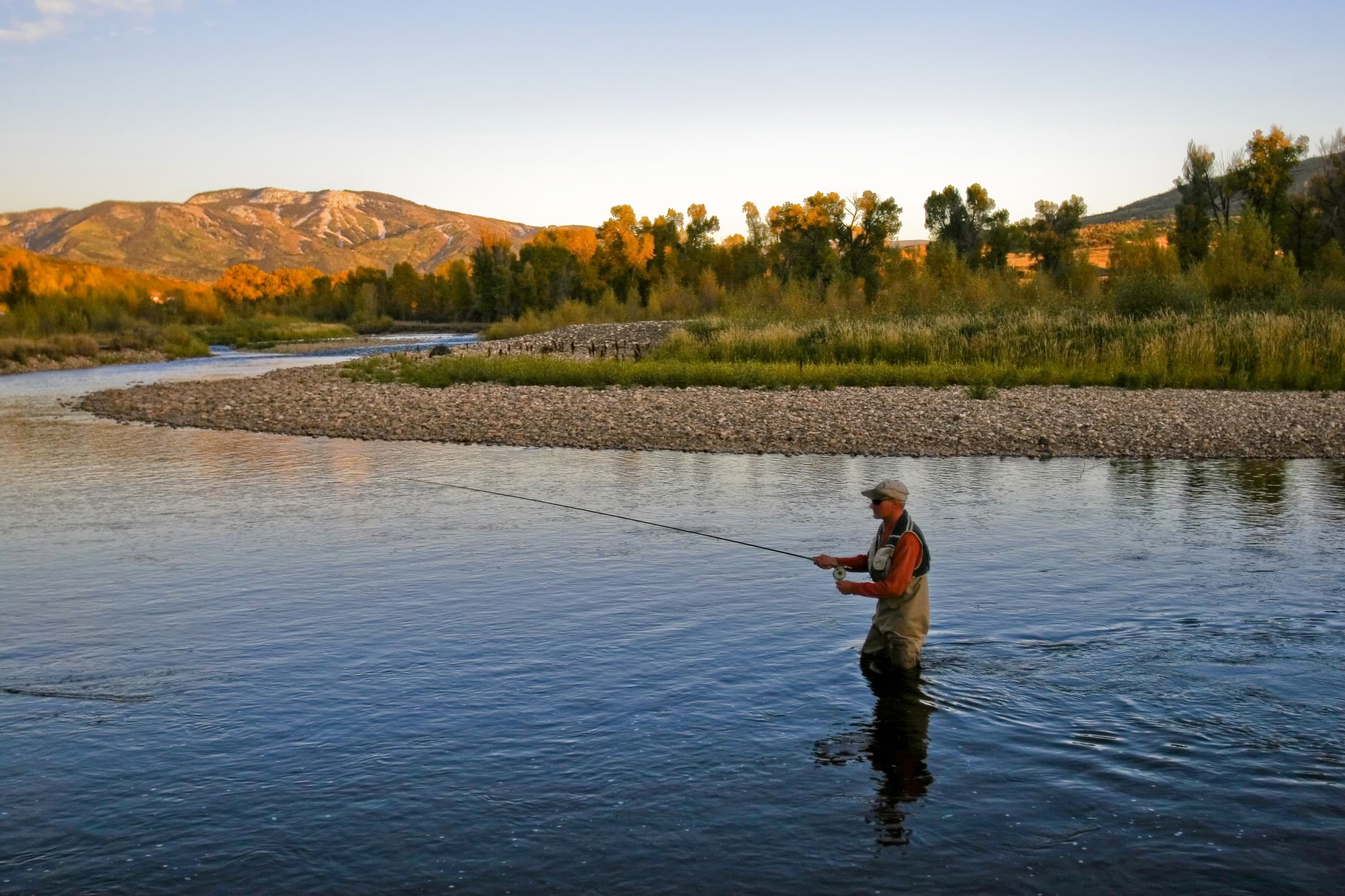 A flyfisher in waders casts his line from the middle of a lake as the sun sets over the mountains in the distance
