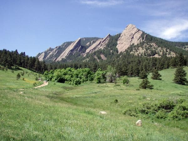Tall slabs of slanted, gray rock are nestled among rolling green foothills and a thick, dark-green forest. The rocks watch over the nearby city of Boulder, Colorado.