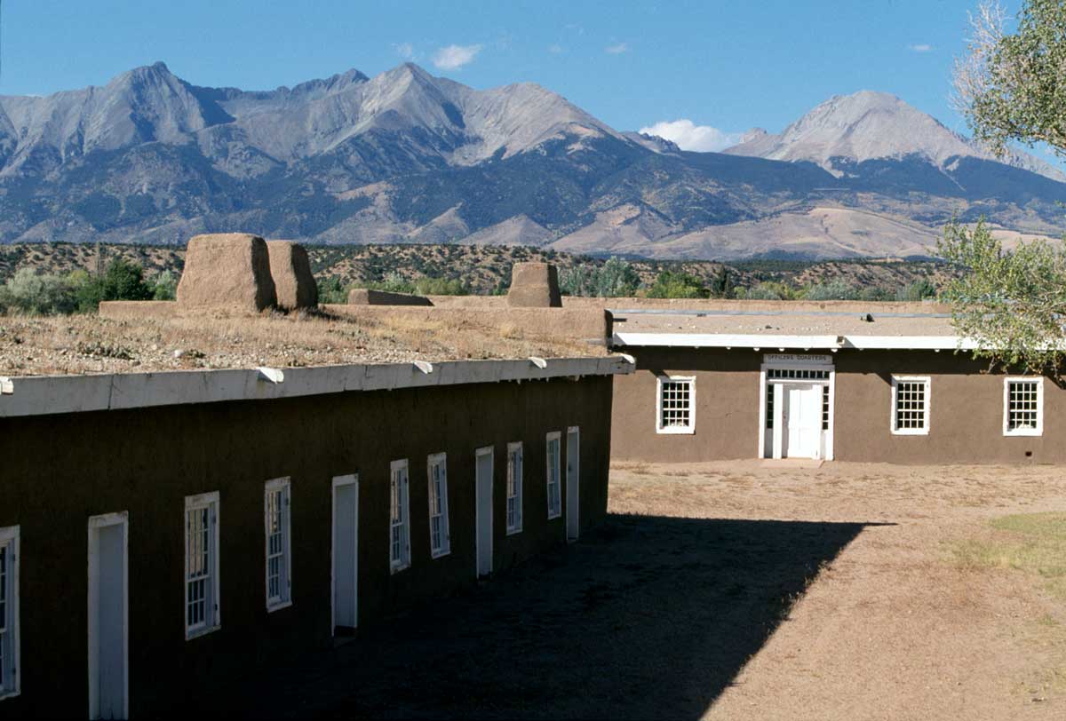 Two small, brown buildings with white-trimmed windows sit on a plot of dirt. These buildings make up the Fort Garland Museum and Cultural Center in Fort Garland, Colorado. Behind the museum is the mighty Sangre de Cristo mountains under a blue sky.