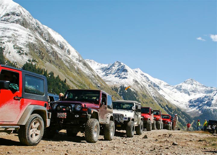 A row of 6 red and white Jeeps with sturdy tires gets ready to brave a rocky road; snowcapped peaks rise in all directions