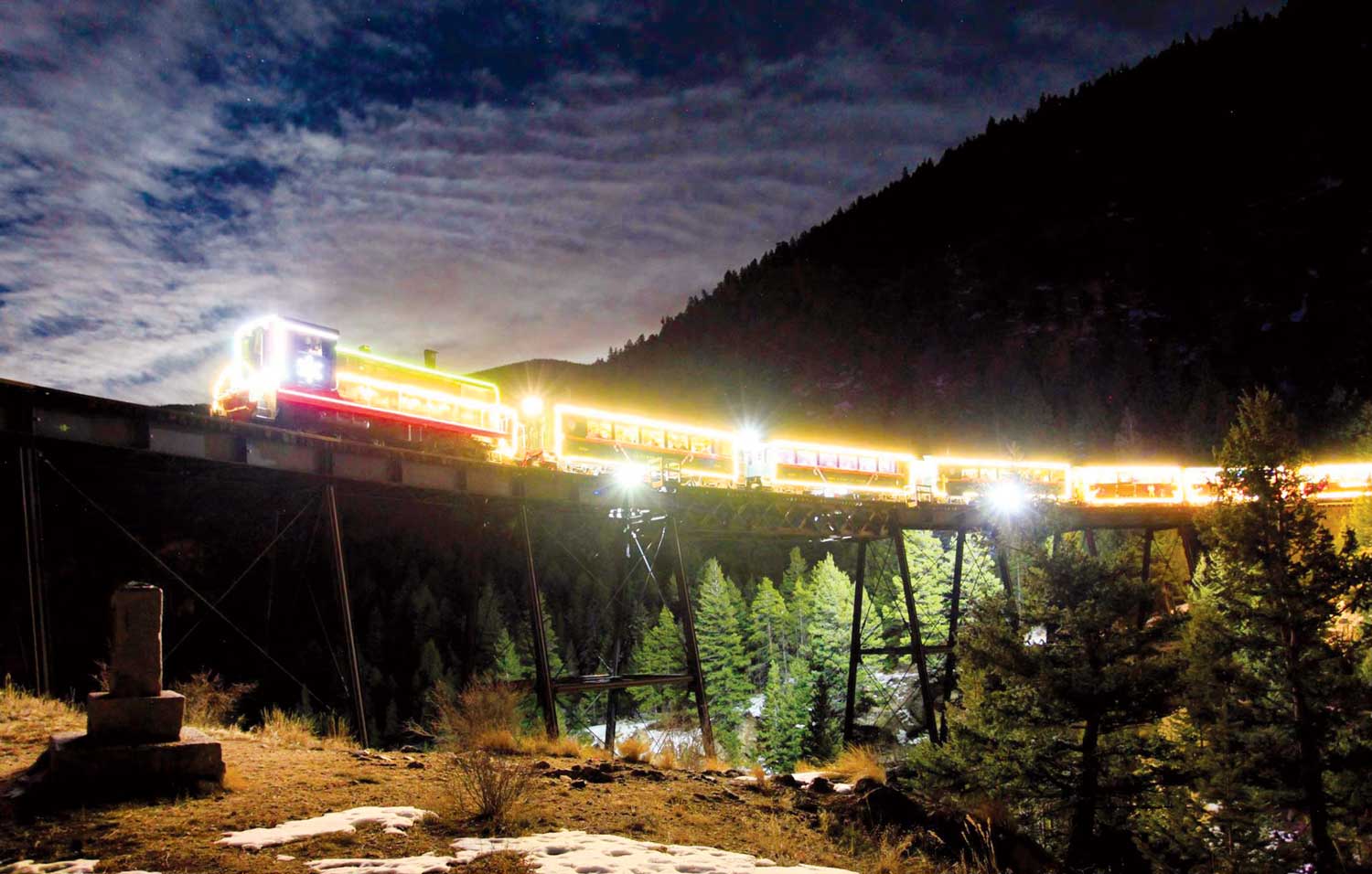 A train glowing with white lights crosses a bridge as the sun sets behind it