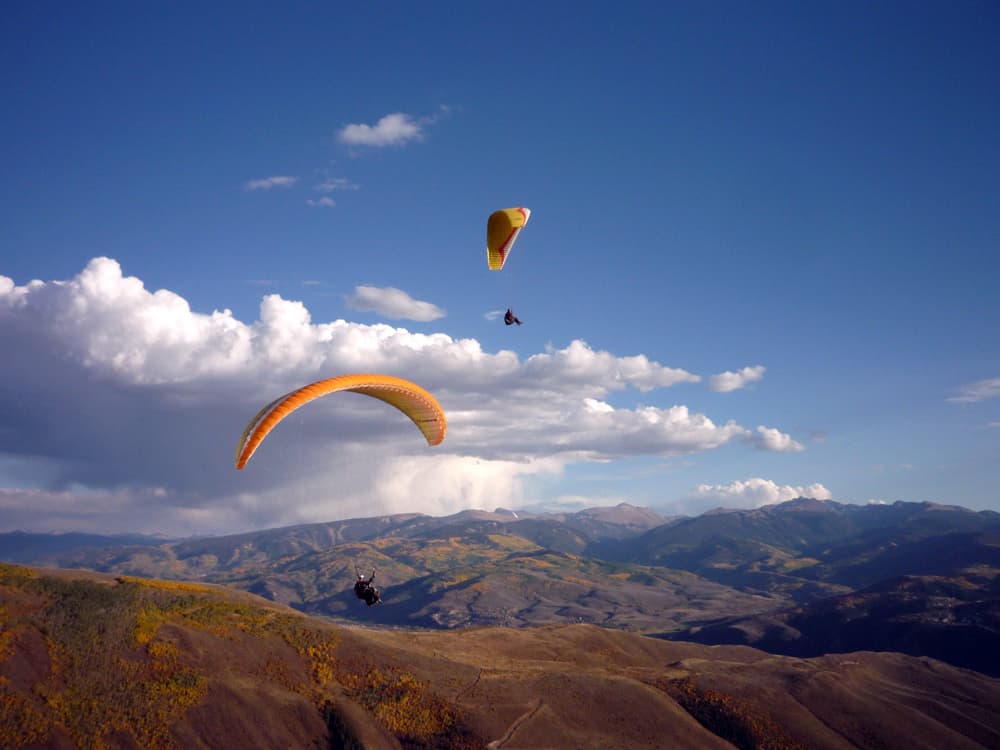 In a mostly blue sky, with a few friendly clouds, we see two paragliders gently moving lower under their orange parachutes into the soft brown foothills of Vail