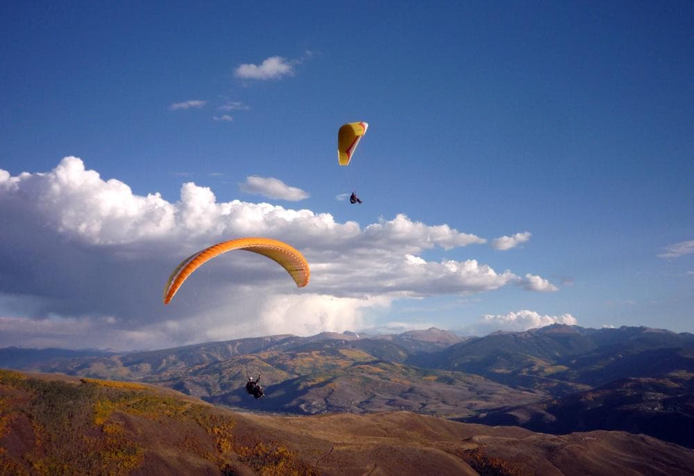 Two hang gliders glide through the blue sky and white clouds over a mountain range in the fall.