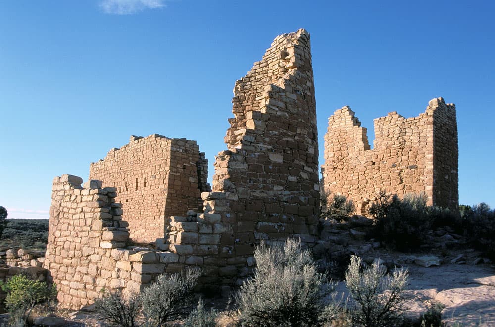 Three stone walled relics sit under a blue sky with scrubby plants at their feet at Hoveweep National Monument.