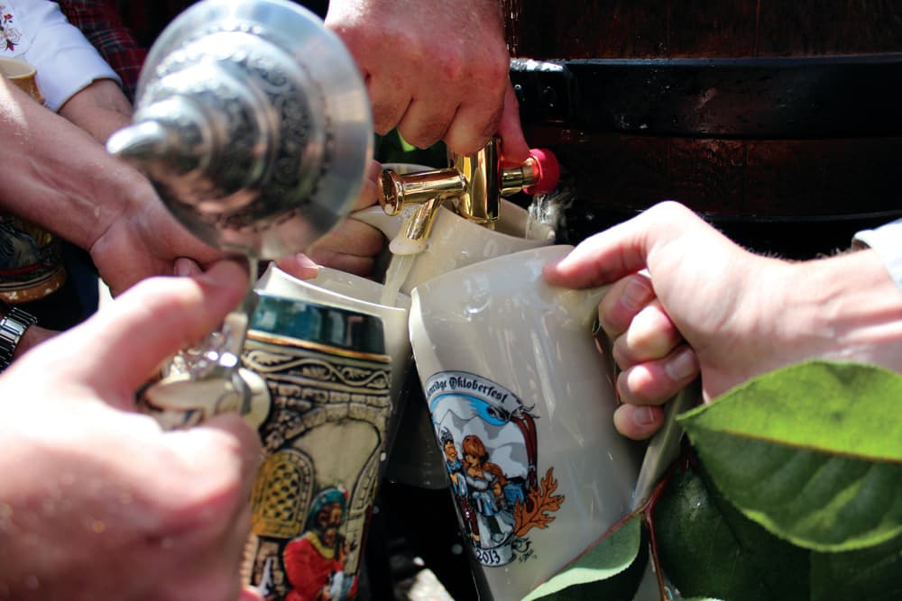 Four decorative Oktoberfest beer steins are positioned around a beer keg with a gold-colored metal spigot waiting to be filled at a Colorado festival.