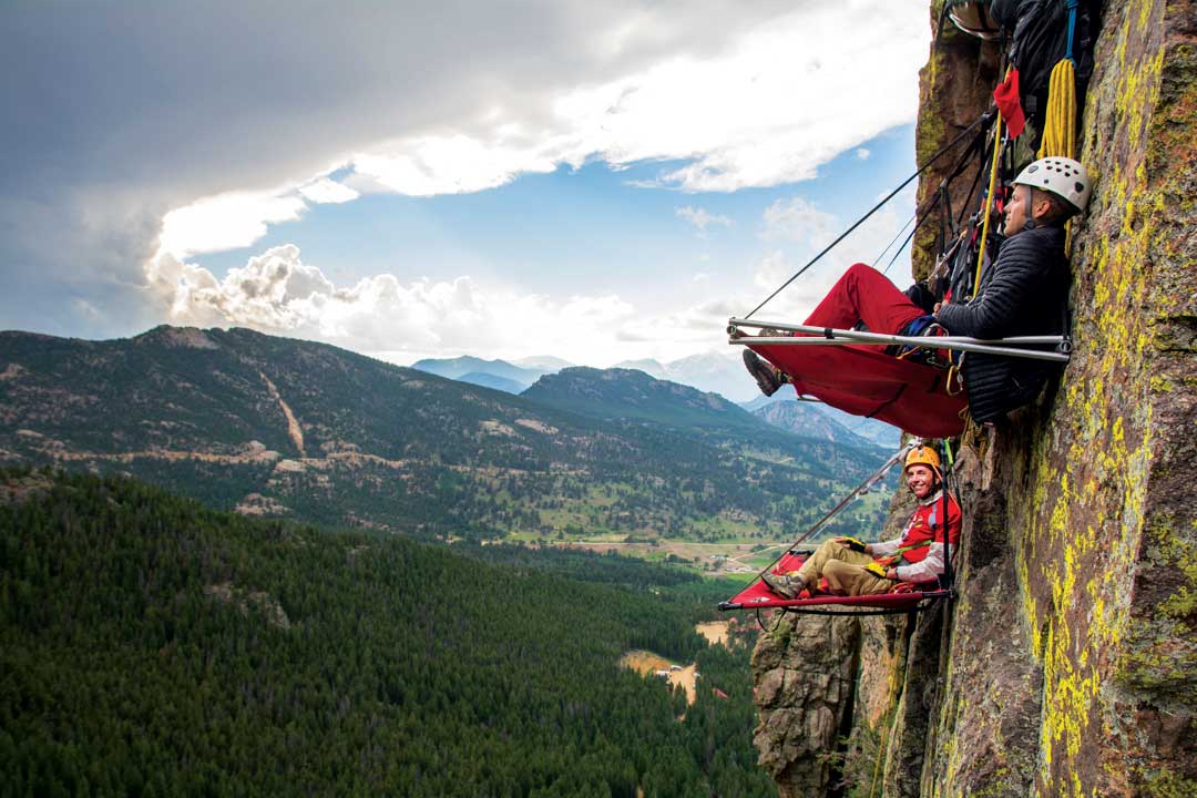 Two climbers sit on red tent-structures with their legs crossed along a nearly-vertical cliff face in Estes Park. They are cliff camping with Kent Mountain Adventure Center and looking out at the magnificent mountainous landscape under a mostly-cloudy sky.