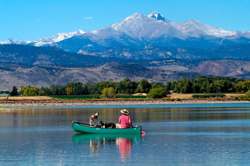 Two curious pups survey the water from a dark-green canoe as the person with them, wearing a red shirt and tan hat, paddles at Union Reservoir in Longmont, Colorado.