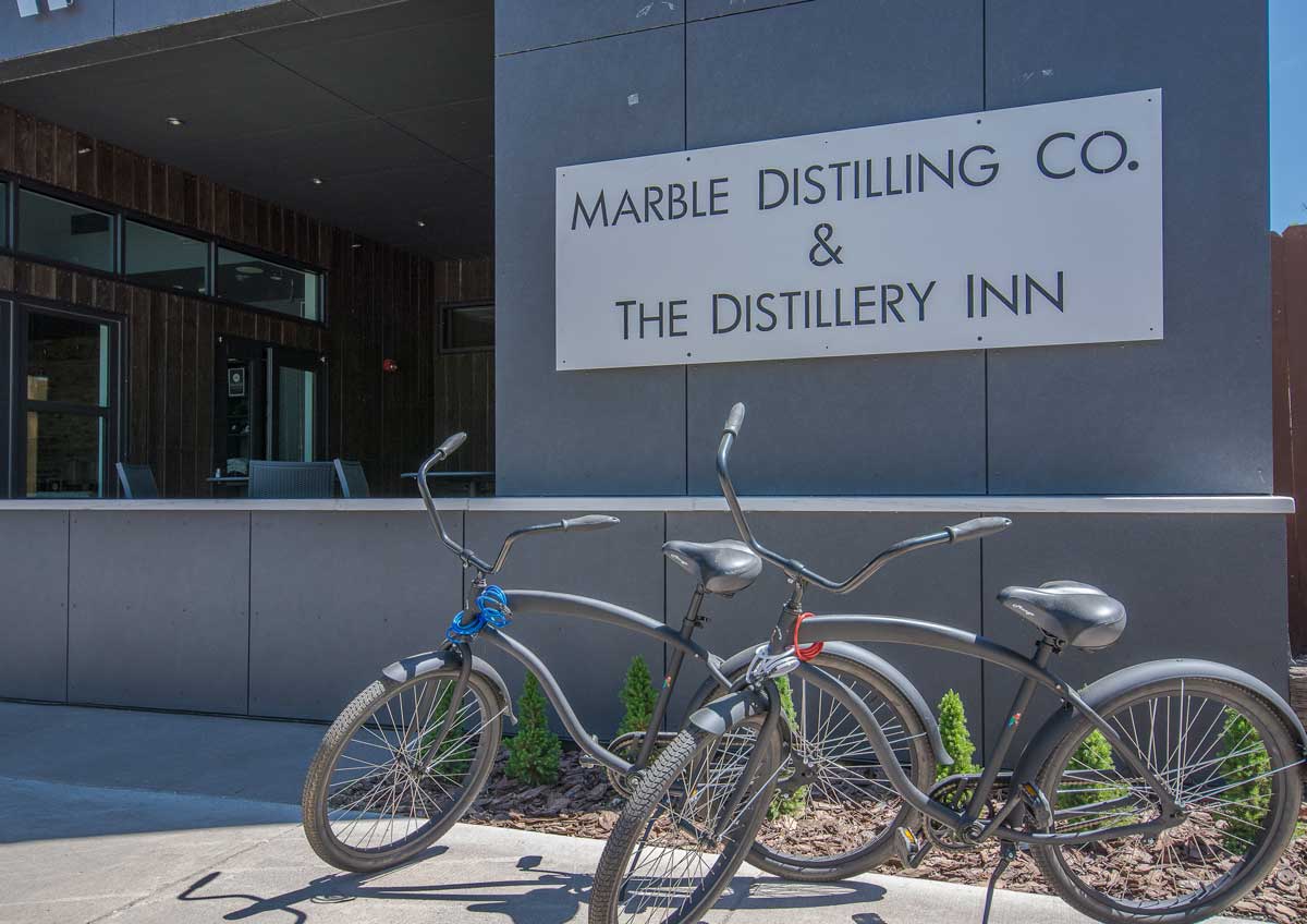 Two gray bicycles with chains wrapped around their frames are parked outside a slate-gray building with a sign for Marble Distilling Co. & The Distillery Inn.