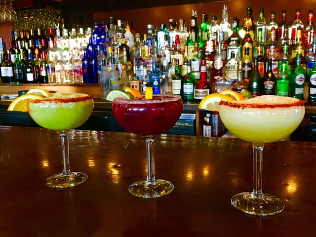 Colorful cocktails sit on a bar with bottles in the background