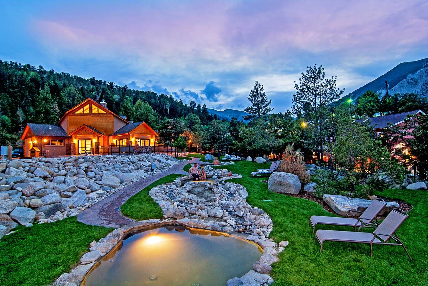 A small pool sits near a dirt path with rocky outcroppings and two pool loungers with a wooden building lit up in the background. There is a mountain ridge with evergreen trees beneath a cloudy, pink-blue setting sun above.