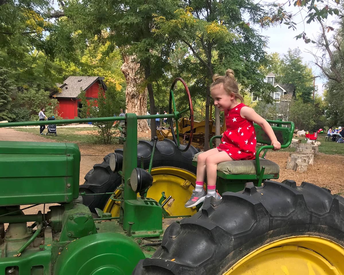 A small child in a red outfit dotted with white stars sits excitedly in the worn, cushioned driver's seat of a older green tractor in Colorado.
