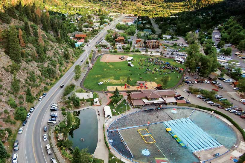 A country highway passes between the Ouray Hot Springs facility and a steep mountainside. The road continues by a crowded park and goes into the town of Ouray.