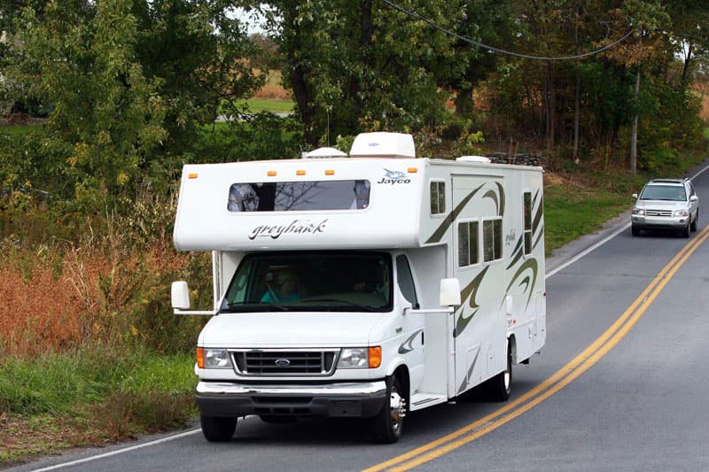 A white RV drives down a leafy road