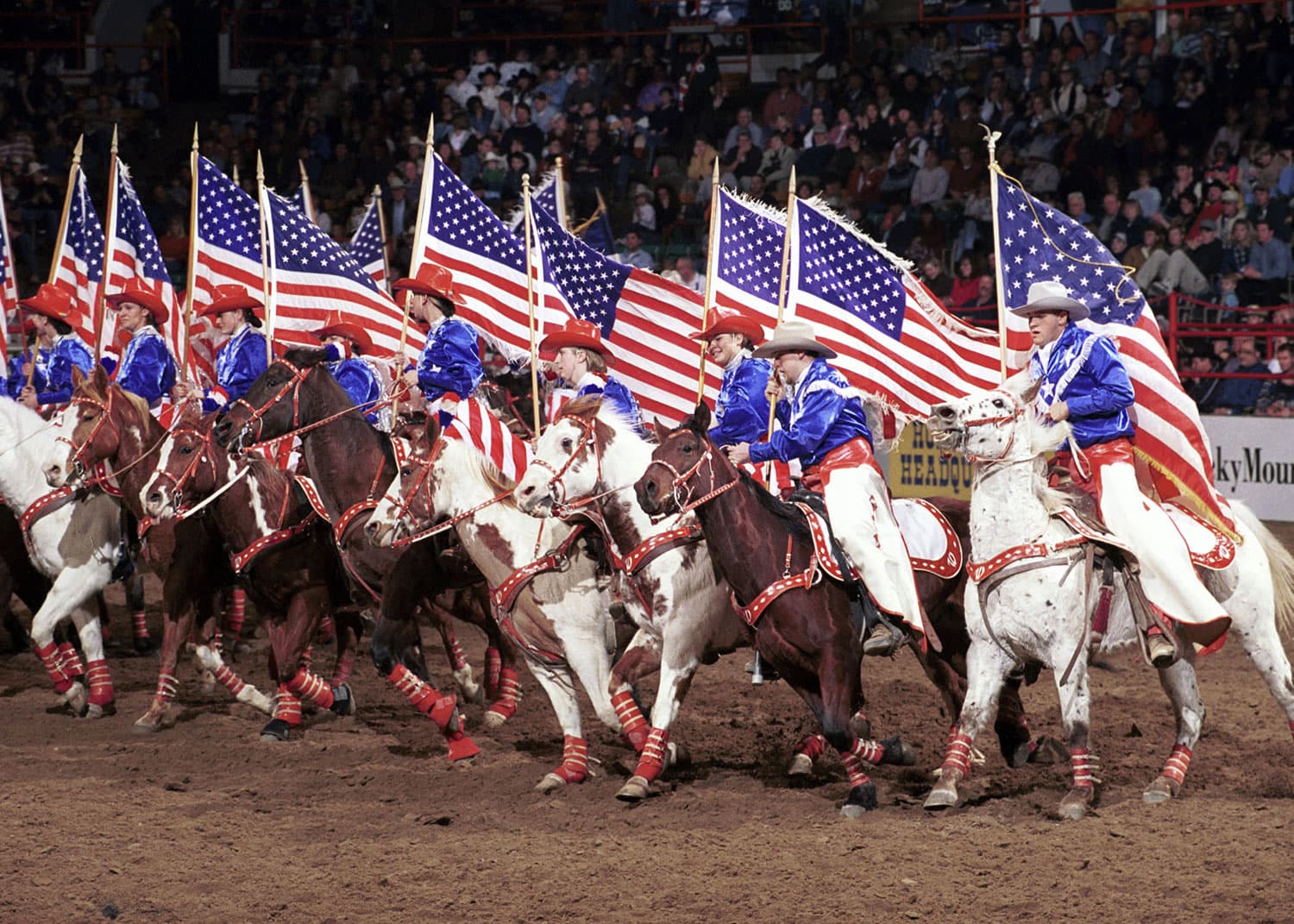 Nine riders on horseback dressed in red-white-and-blue outfits holding waving American flags race through the dirt of the stadium at the National Western Complex in Denver. This group of horse riders is the Westernaires, performing at National Western Stock Show.