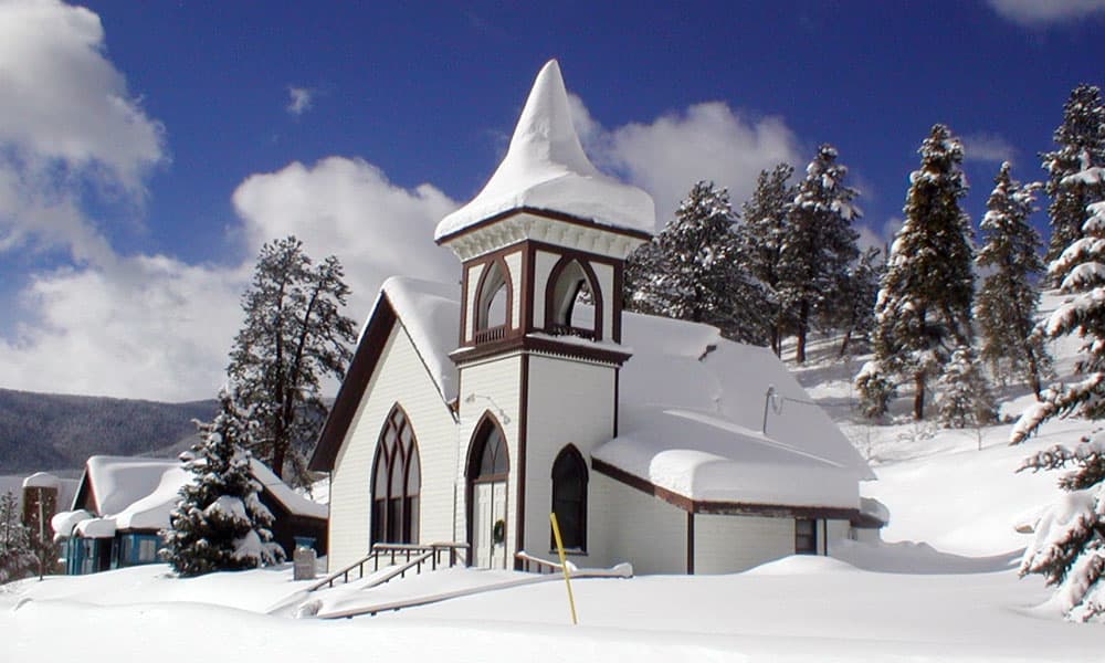 Bright-white snow blankets the historical Pitkin Community Church in downtown. There's a blue sky with white clouds and evergreen trees.